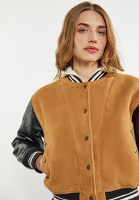 Giubbotto bomber in suede marrone con maniche in pelle nera, bottoni a pressione, polsini e orlo a coste, e una felpa a righe che spunta dal colletto.