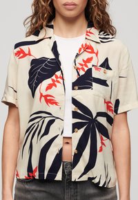 Camisa de manga corta en color crema con patrones de hojas tropicales en rojo y azul marino, que incluye un bolsillo en el pecho y botones redondos marrones. Tejido texturizado.