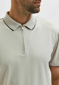 Polo shirt gris clair avec un col texturé, doté d'une fermeture éclair demi-longueur et de détails noirs sur le bord du col. Tissu doux et lisse.