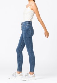 Jeans azules de cintura alta y corte ajustado con botones, llevados por una persona en una camiseta sin mangas blanca y zapatillas blancas, de pie en perfil lateral.