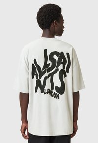Persona vestida con una camiseta blanca de gran tamaño con un texto negro distorsionado que dice "ALLSAINTS LONDON" en la parte de atrás, de espaldas.