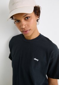 Svart bomulls T-skjorte med en hvit "obey" logo på venstre side, kombinert med en lys beige, brodert caps. Myk tekstur, avslappet passform.
