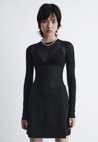 Schwarzes langärmliges Oberteil aus durchsichtiger Stoff, das über einen enganliegenden schwarzen Rock getragen wird. Hat einen Herzausschnitt und eine silberne Kettenhalskette.