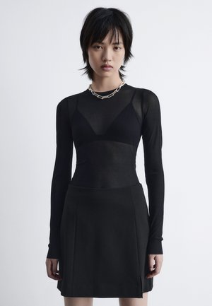 Black long-sleeve top in sheer fabric layered over a fitted black skirt. Features a sweetheart neckline and a silver chain necklace.