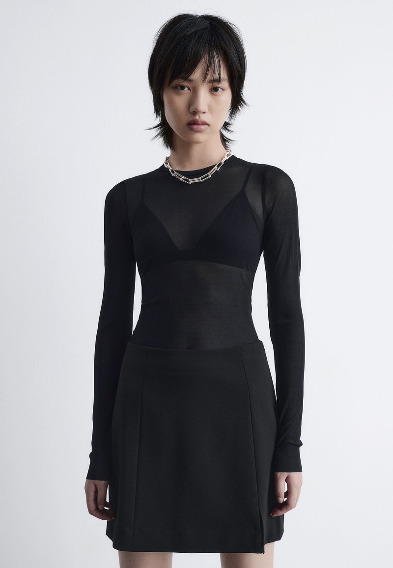 Schwarzes langärmliges Oberteil aus durchsichtiger Stoff, das über einen enganliegenden schwarzen Rock getragen wird. Hat einen Herzausschnitt und eine silberne Kettenhalskette.