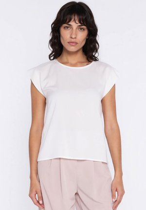 Kobieta o ciemnych falowanych włosach, ubrana w biały, bezrękawnik i jasnobeżowe spodnie, stojąca na tle jednolitego białego tła.