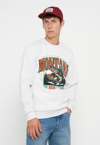 Jeune homme portant un sweat-shirt blanc avec un graphique de pêche coloré du Montana et une casquette de baseball rouge, se tenant devant un fond uni.