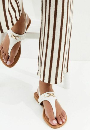 Pieds chaussés de sandales blanches à lanières avec des détails de boucle dorée, assorties à un pantalon crème avec des rayures verticales marron.