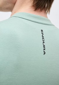 Κοντινή λήψη του πάνω μέρους της πλάτης ενός ατόμου που φοράει ένα ανοιχτό πράσινο πουκάμισο με κάθετο μαύρο λογότυπο "ENDURA" κοντά στον γιακά.