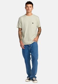 Maglietta di cotone verde chiaro con collo rotondo e maniche corte, con taschino sul lato sinistro del petto e logo nero. Indossata con jeans blu.
