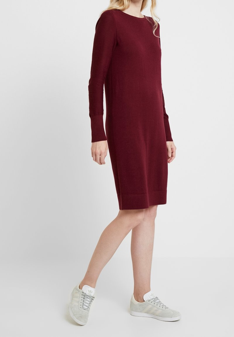 Femme portant une robe burgonde à manches longues de longueur genoux et des baskets Adidas gris clair, se tenant devant un fond blanc uni.