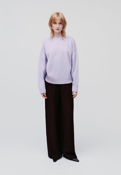 Lila oversized trui met ribbels aan de mouwen, gecombineerd met wijde zwarte broek en puntige schoenen, tegen een egale witte achtergrond.