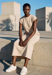 Young woman in beige dress and white sneakers sitting on concrete ledge outdoors with large concrete blocks and palm shadows in sunlight.