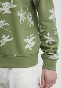 Groene sweatshirt met een wit loofpatroon, geribbelde manchetten en zoom, zachte textuur, met een merketiket aan de zijkant. Joggingbroek in grijs.