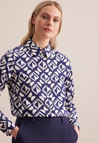 Camicia a maniche lunghe blu e bianca con motivi geometrici, dotata di colletto e bottoni sul davanti. Il tessuto appare liscio con una vestibilità strutturata.