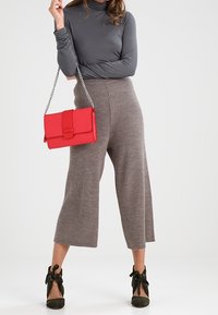 Haut gris à col roulé associé à un pantalon beige large et côtelé. Un sac à main rouge vif avec un fermoir, une chaîne argentée, et des bottines vert foncé avec des nœuds.