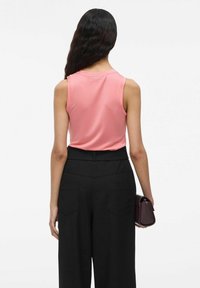 Ärmlös rosa topp, svarta högmidjade byxor, texturerat hår, håller en liten vinröd handväska. Enkel, avslappnad outfit, sedd bakifrån.