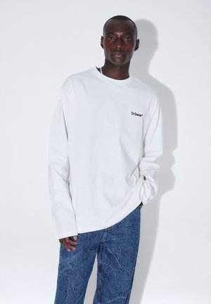 Mand iført en hvid langærmet skjorte med teksten "Dr. Denim" og strukturerede blå jeans, stående mod en ensfarvet hvid baggrund.