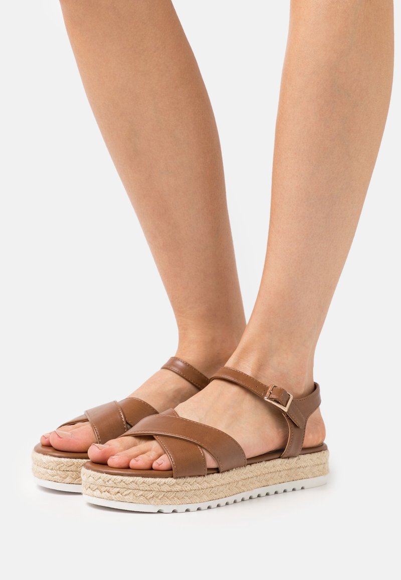 Brown leather sandals with crossed straps, adjustable ankle buckle, and espadrille-style woven platform, featuring a white rubber sole.