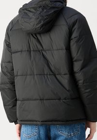 Czarna pikowana kurtka puchowa z kapturem noszona przez osobę w niebieskich dżinsach, widoczna z tyłu na jednolitym jasnym tle.