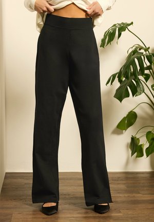 Persoon die een crèmekleurige trui omhoog trekt, draagt zwarte broek met wijde pijpen en zwarte puntige schoenen, staand op een houten vloer nabij een groene plant.