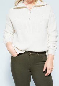 Suéter blanco con textura y media cremallera, con detalles en canalé y aberturas laterales, combinado con unos jeans ajustados de color oliva que tienen bolsillos frontales y herrajes dorados.