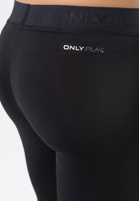 Leggings neri realizzati in materiale elasticizzato, con una vita alta dotata del logo "ONLY PLAY" centrato in bianco, texture liscia e design aderente.