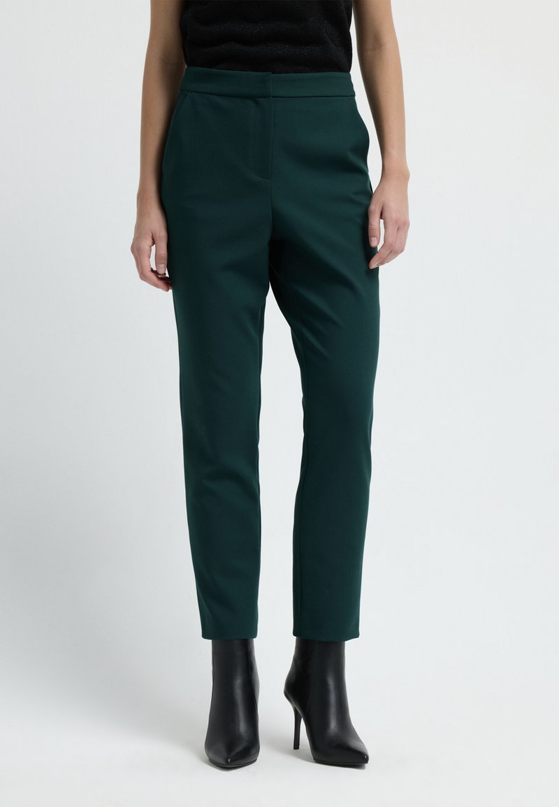 Pantaloni su misura verde scuro con vestibilità slim, realizzati in un tessuto liscio. Presentano un alto punto vita e tasche laterali, abbinati a stivali neri con tacco.
