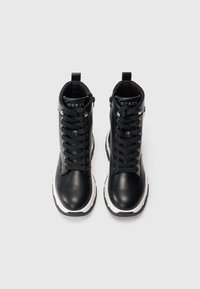 O pereche de adidași negri cu glezna înaltă, cu tălpi groase albe și șireturi negre, marca "GUESS" pe limbă căptușită, prezentată din vedere de sus pe un fundal alb.
