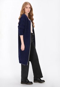 Hosszú sötétkék kardigán texturált kötött anyagból, nyitott elülső részével és kontrasztos világos díszítéssel. Egy szűk felső és fekete nadrág felett viselve.