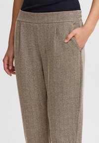 Bruine broek met een visgraatpatroon, voorzien van een elastische tailleband en zijzakken. Zacht materiaal met een relaxte pasvorm en subtiele textuur.