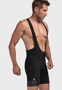 Schwarze Radträgerhose mit suspendierten Trägern, flachen Nähten, enger Passform und einer Seitentasche. Reflektierendes Logo am Unterschenkel.