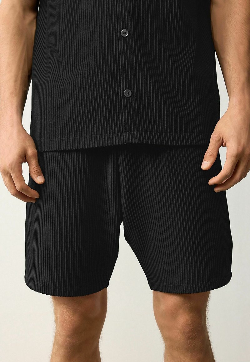Chemise noire à rayures verticales à manches courtes et shorts assortis. Les deux vêtements présentent une texture côtelée verticale et des boutons sur la chemise.