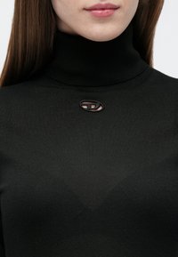 Γυναίκα που φοράει μαύρο πουλόβερ με ζιβάγκο, με ένα μικρό οβάλ άνοιγμα και διακοσμητικό κούμπωμα κάτω από το γιακά.