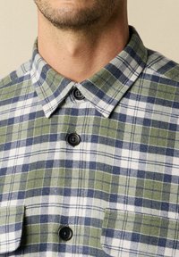 Camicia a quadri verde e blu con colletto abbottonato, dotata di due tasche frontali e bottoni neri. Realizzata in tessuto intrecciato con motivo a quadri.