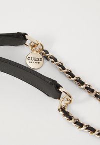 Svart faux skinn handbagreim med gullfargede metallakenter og en dekorativ kjede. Har et rundt merketag med "GUESS EST.1981."