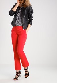 Chaqueta de cuero negra, top con estampado de pata de gallo, pantalones rojos brillantes y tacones negros con tiras. La modelo posa con una mano en la cadera.