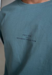 Teal katoenen T-shirt met korte mouwen, met "PHIL & CO. WEEKEND COLLECTION" bedrukt in donkerblauw op de borst. Glad aanvoelend.