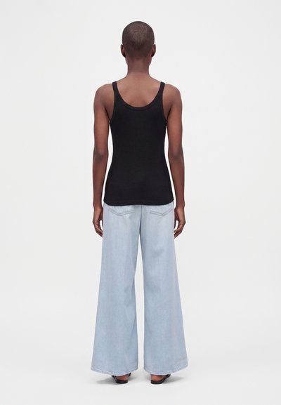 Personne aux cheveux courts portant un débardeur noir et un jean large bleu clair, debout, dos tourné, sur un fond uni.