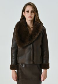 Giacca di pelle marrone con un ampio colletto e polsini in pelliccia sintetica. Texture liscia con un design aderente. Il modello indossa una gonna abbinata con motivo coccodrillo.