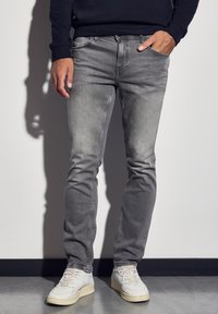 Jeans de denim gris con un acabado desgastado, corte slim, diseño de cinco bolsillos, combinados con zapatillas blancas, de pie frente a un fondo neutro.