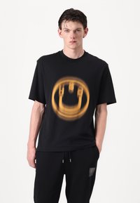 Sort bomulds t-shirt med et stort orange grafisk print af et smilende ansigt på fronten. Korte ermer og rund halsudskæring.