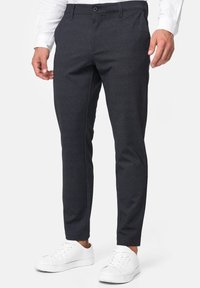 Hombre vistiendo pantalones oscuros ajustados con botón en la parte frontal y bolsillos, combinados con zapatillas blancas y una camisa de manga larga blanca.