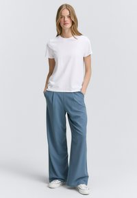 Witte korte mouwen t-shirt gecombineerd met losse, lichtblauwe wijde broek. Model staat met de handen in de zakken, met witte sneakers aan.