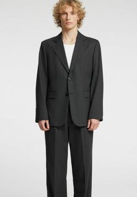 Completo grigio scuro su misura con blazer strutturato, chiusura a un bottone e pantaloni a vita piatta. Il tessuto appare liscio, con un leggero lucido.