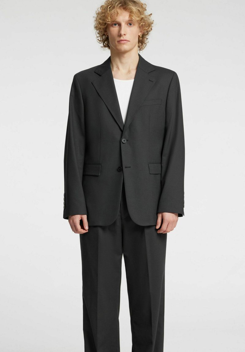 Completo grigio scuro su misura con blazer strutturato, chiusura a un bottone e pantaloni a vita piatta. Il tessuto appare liscio, con un leggero lucido.