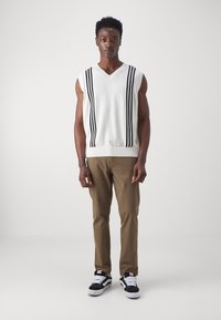White sleeveless sweater with black vertical stripes, paired with brown pants and black sneakers with white accents. Model stands upright.