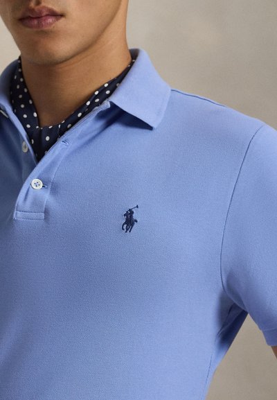 Polo azul claro com textura canelada, apresenta uma pala com dois botões e um pequeno logótipo escuro no lado esquerdo do peito. Acompanha um lenço escuro com bolinhas.