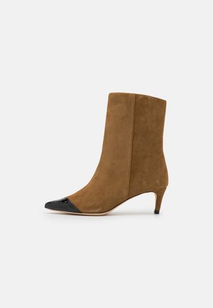 Botte mi-mollet en daim marron avec un bout pointu noir brillant et un petit talon kitten sur un fond gris.