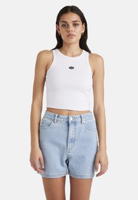 Weißes, geripptes Croptop mit schwarzem Logo auf der Brust, kombiniert mit hochgeschnittenen, hellblauen Jeansshorts mit vorderen Taschen und Knopfverschluss.
